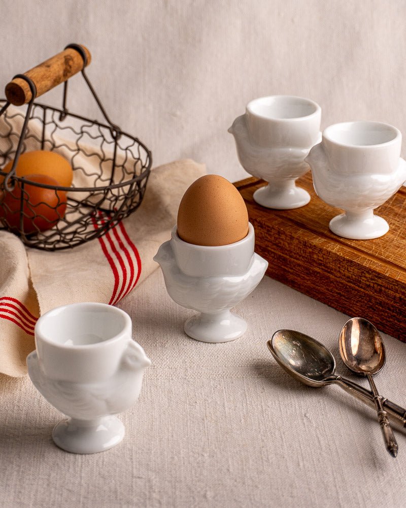 White vintage French Opaline glass egg cup with brown egg, coquetiers and eggs in background.