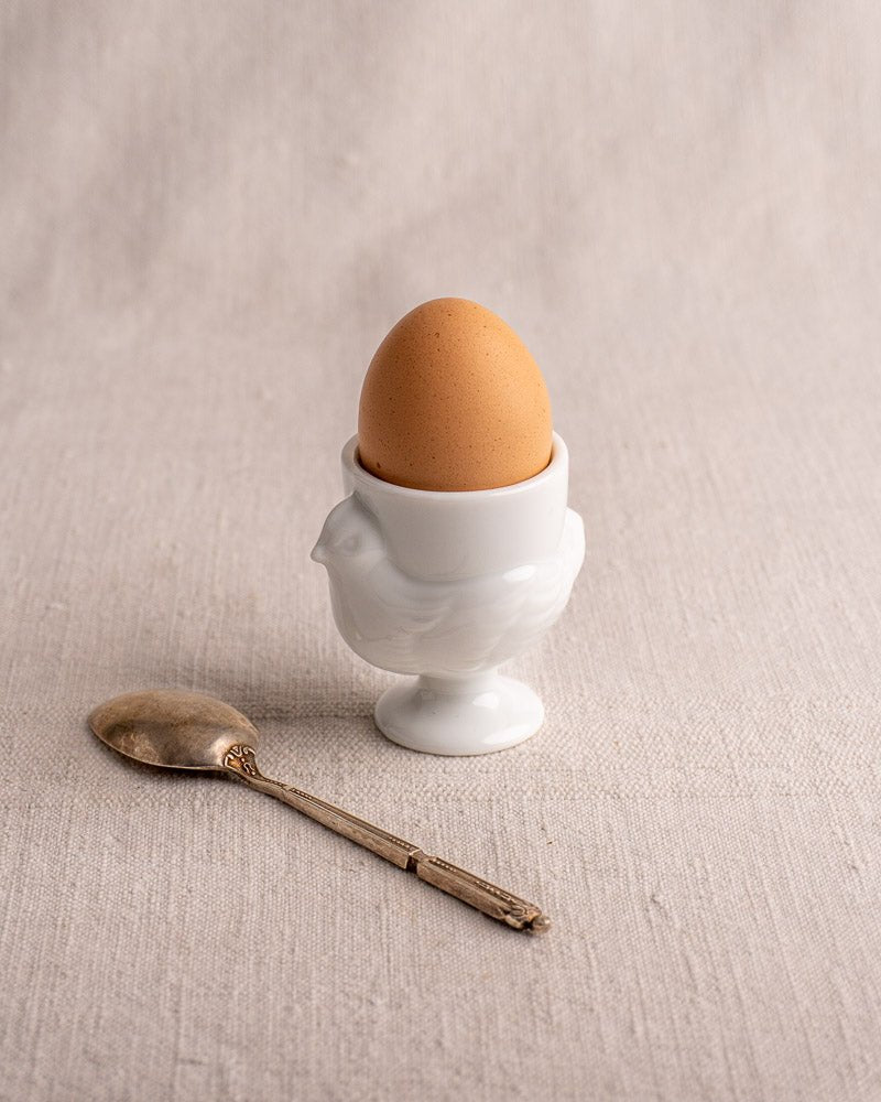 Vintage French opaline glass hen coquetier with brown egg and antique silver spoon on beige textured surface.