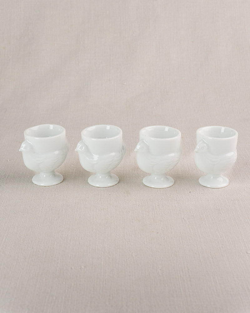 Four vintage French opaline glass hen-shaped egg cups arranged on beige fabric.