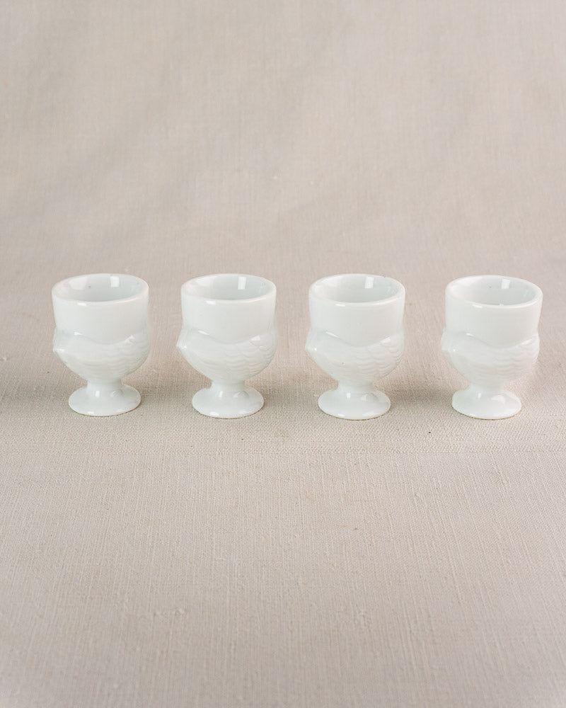 Set of four vintage French opaline glass hen-shaped egg cups on beige fabric surface.