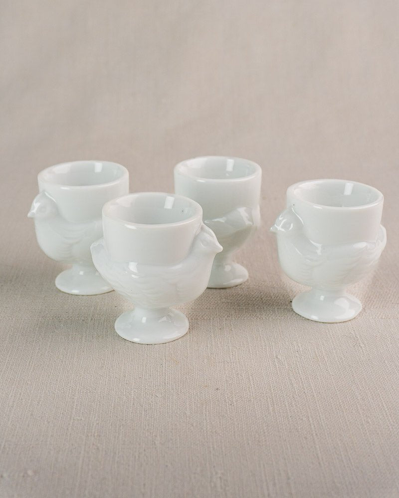Set of four vintage French opaline glass egg cups shaped as hens on a neutral fabric surface.