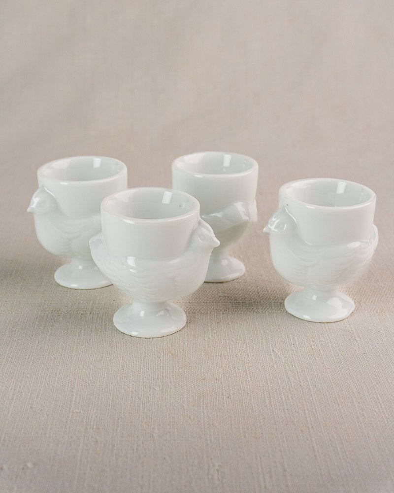 Set of four vintage French opaline glass hen-shaped egg cups on light beige fabric.