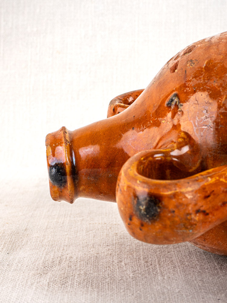 Antique French glazed terracotta wine gourde with two handles, orange-brown ceramic on beige fabric.