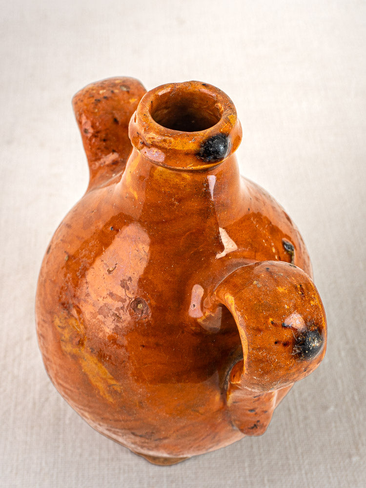 Antique French terracotta wine gourde with orange-brown glaze, spout, and two large handles on light textured surface.