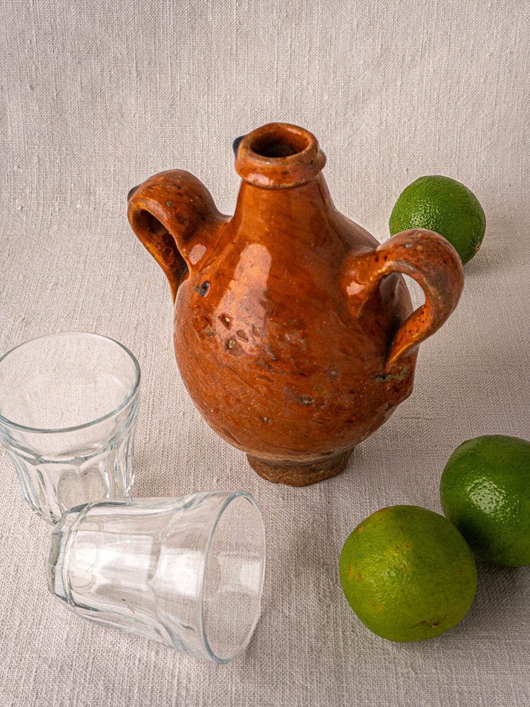 Antique French terracotta wine gourde with two handles, styled with glasses and limes on textured cloth.