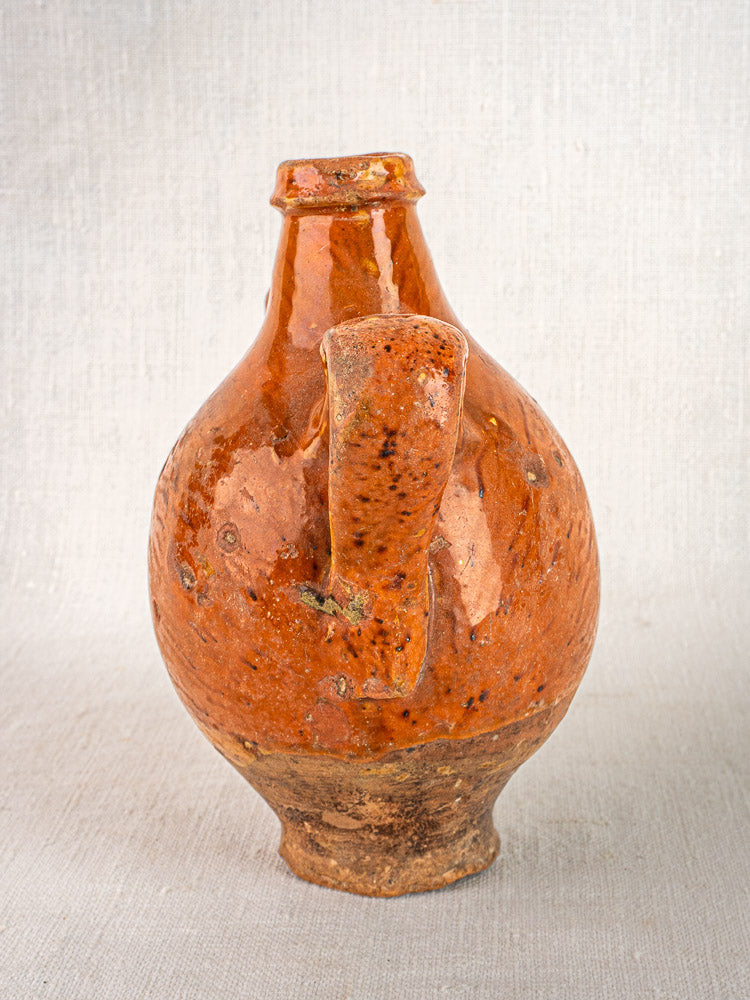 Antique French glazed terracotta wine gourde with two handles and tilted neck, on light textured surface.