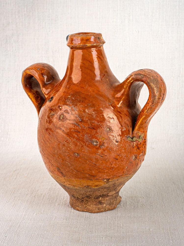 Antique French terracotta wine gourde with rustic brown glaze, two handles, on neutral fabric background.
