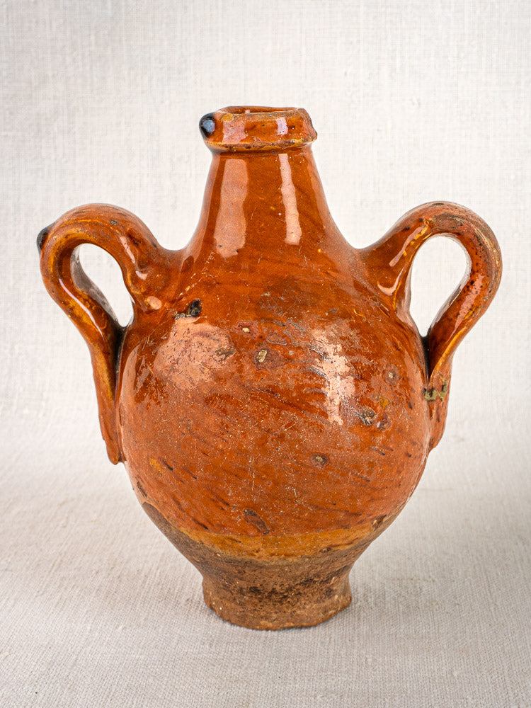 Round brown glazed terracotta French wine jug with two handles and short wide neck on light fabric.