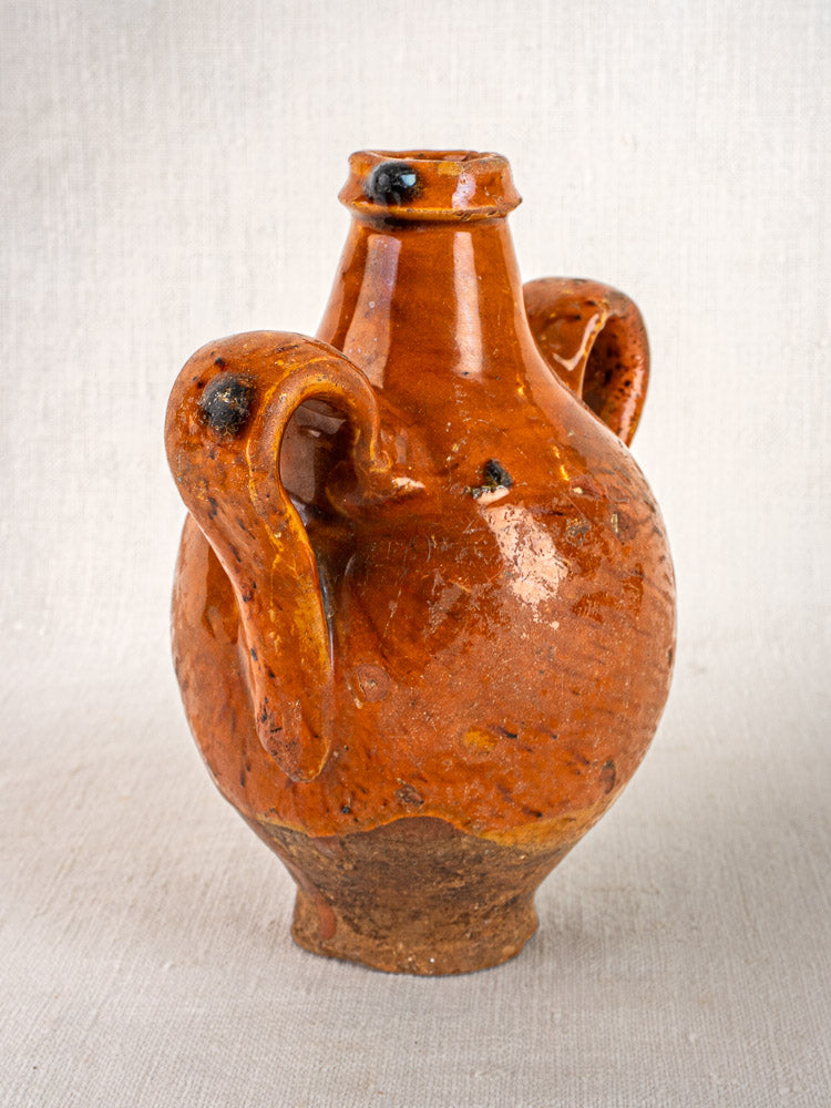 Antique French glazed terracotta wine gourde with two curved handles on a neutral fabric backdrop.