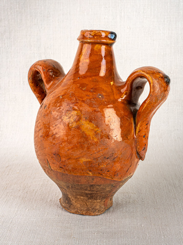 Antique French terracotta wine gourde with rustic brown glaze, two handles, and narrow neck on fabric.