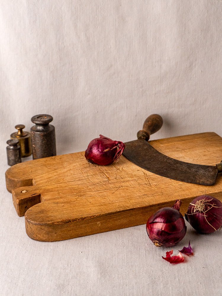 Antique French double-lobed hardwood cutting board with curved knife, red onions, and metal weights on neutral fabric.
