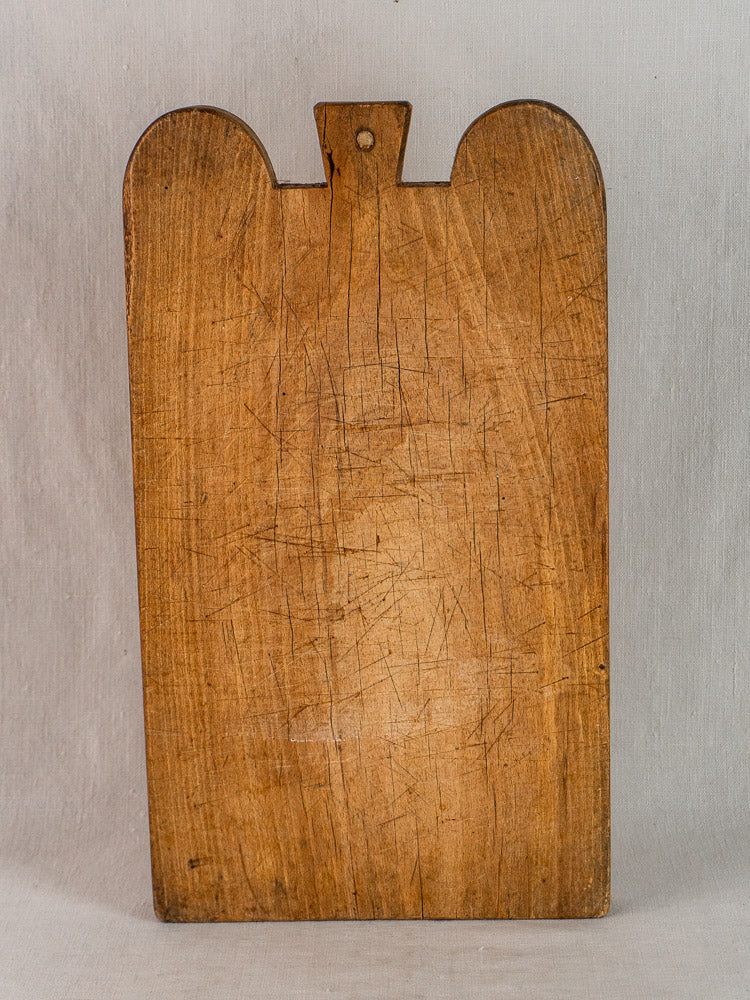 Antique French hardwood cutting board with double-lobed shape, worn surface, and hanging hole on gray background.