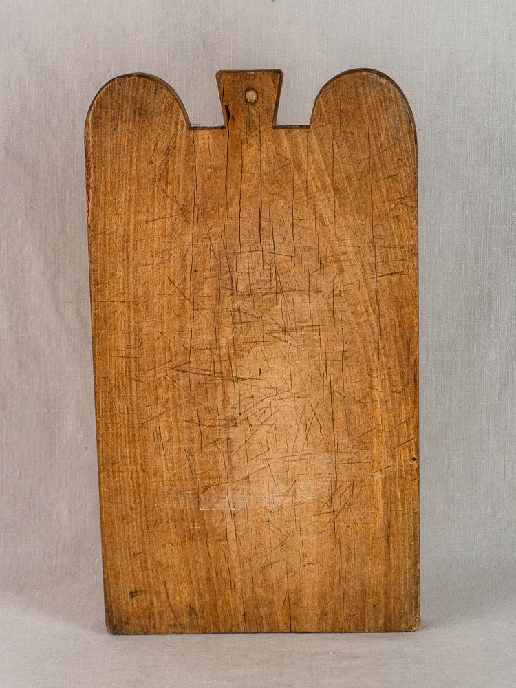 Antique French hardwood cutting board with double-lobed shape, worn surface, and hanging hole on gray background.