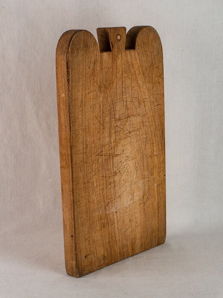 Antique French double-lobed hardwood cutting board with knife marks, upright against a neutral background.