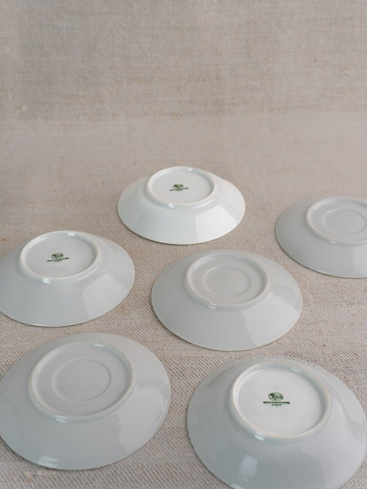 Six white porcelain Hutschenreuther saucers, upside down, display green logos on neutral fabric.