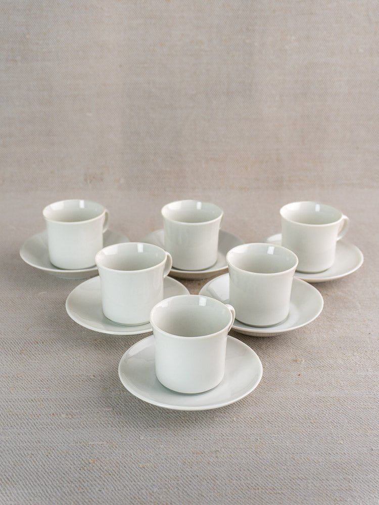 Set of six white porcelain Hutschenreuther espresso cups with saucers arranged on a textured beige surface.