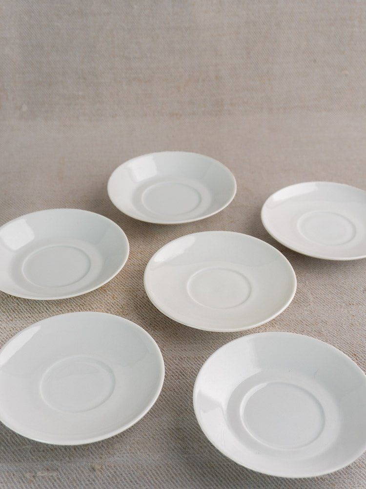 Set of six white Hutschenreuther porcelain saucers in loose circular arrangement on beige textured fabric.