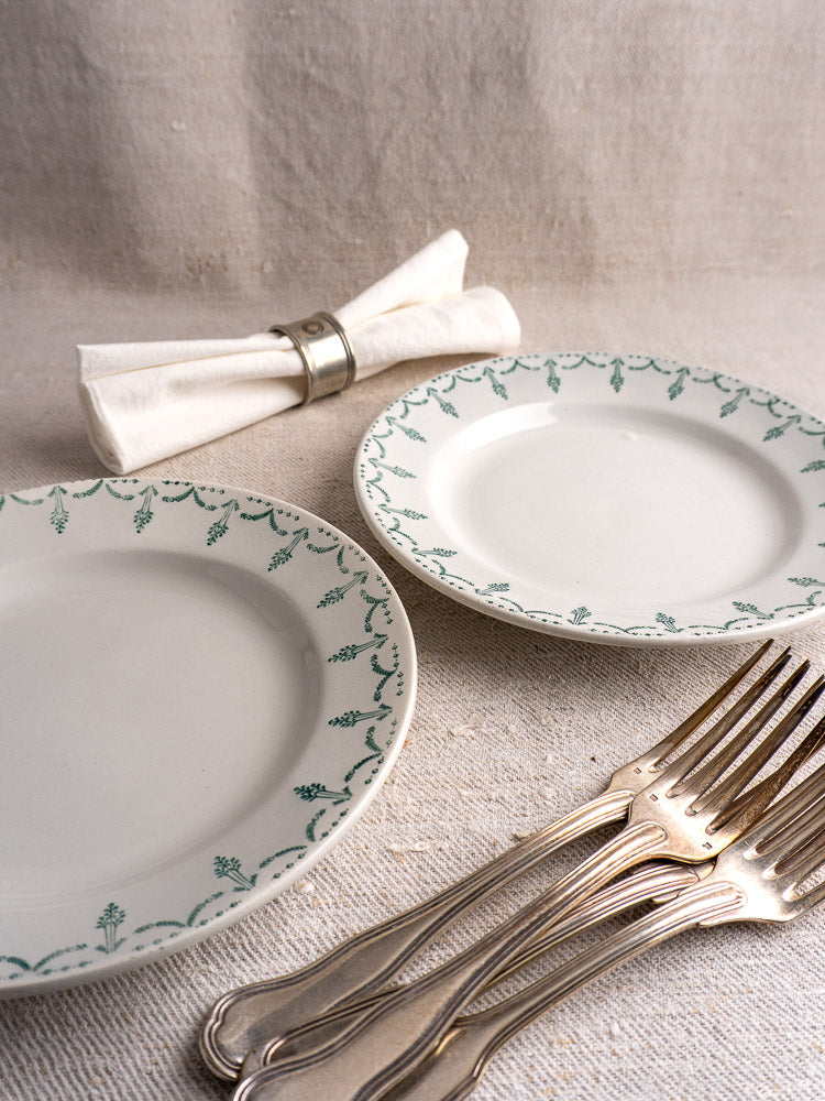 Pair of antique French Gien porcelain plates with green garland border, silver forks, and napkin on beige surface.