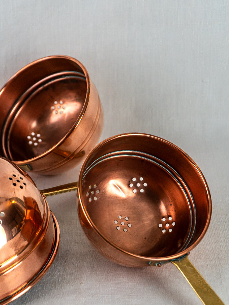 Antique French copper passoire set with brass handles and floral perforations on a light gray surface.