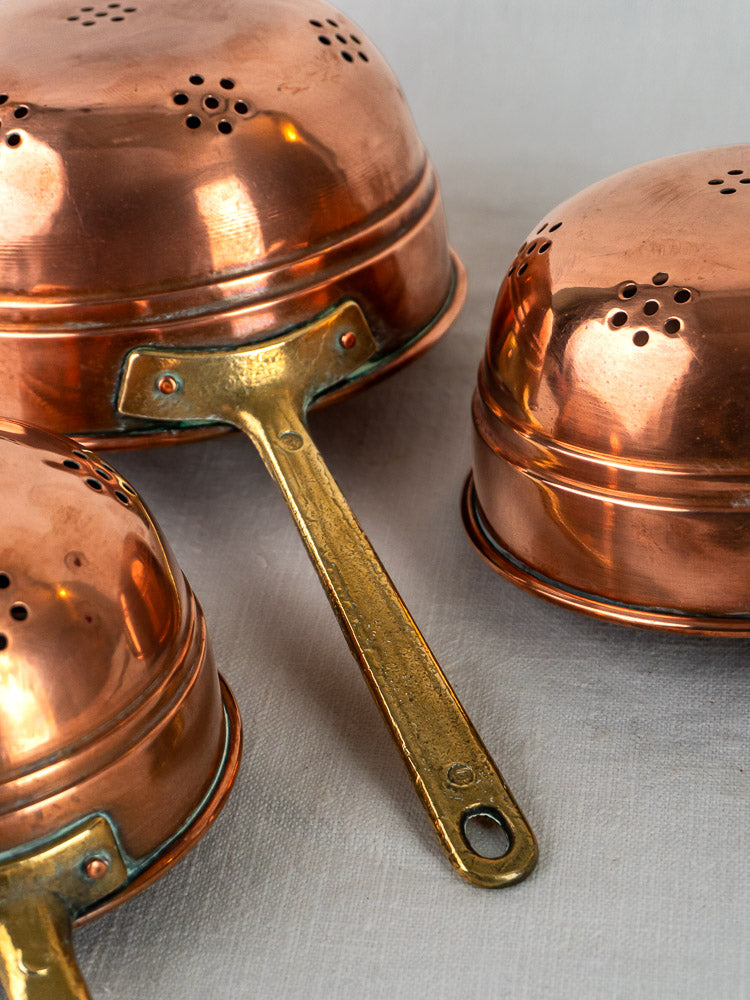 Antique French copper colander trio with brass handles and perforations on a light surface.