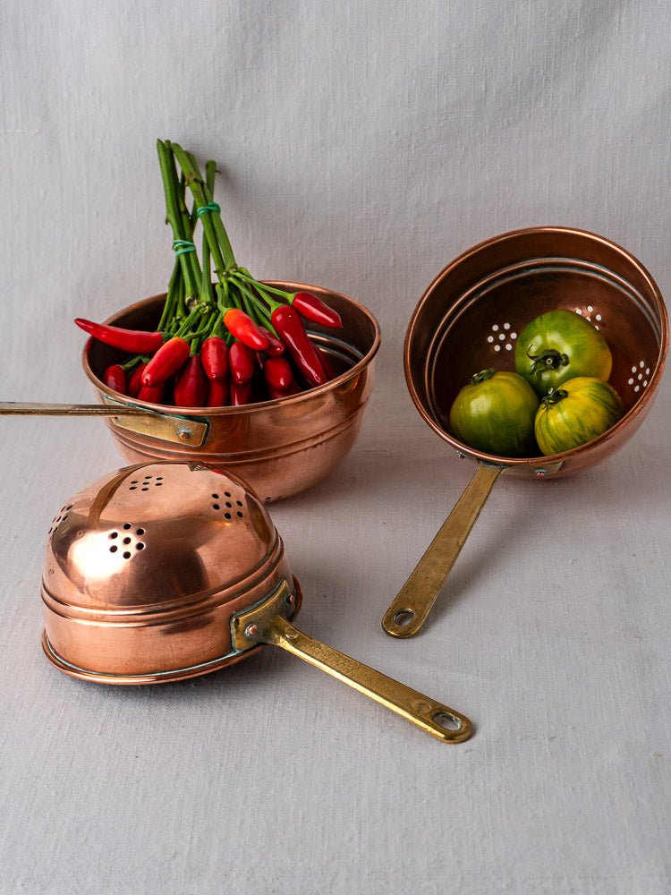 Antique French copper colander trio with brass handles, showcasing perforated patterns on a neutral background.