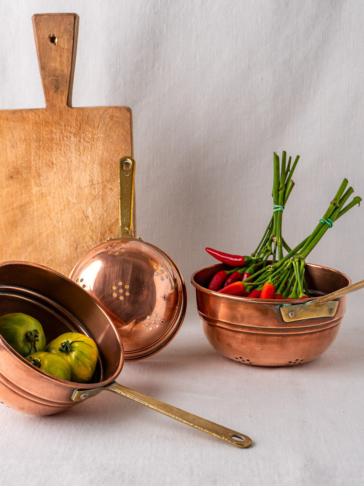 Antique French copper passoire colanders filled with green tomatoes and red chili peppers, wooden board behind.