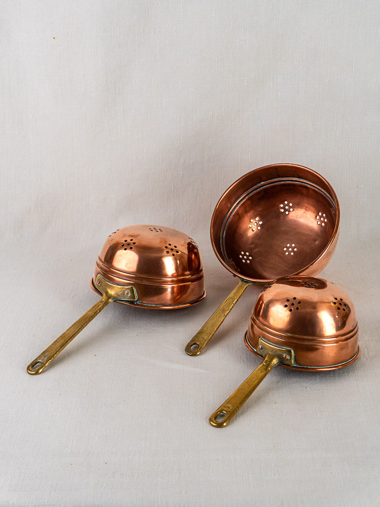 Antique French copper colander trio with brass handles, showcasing perforated patterns on a neutral background.