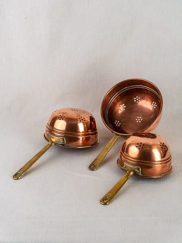 Antique French copper colander trio with brass handles, showcasing perforated patterns on a neutral background.