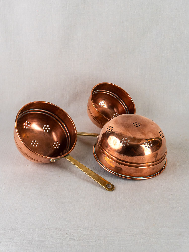 Antique French copper passoire colander trio with brass handles, nested on a light fabric background.