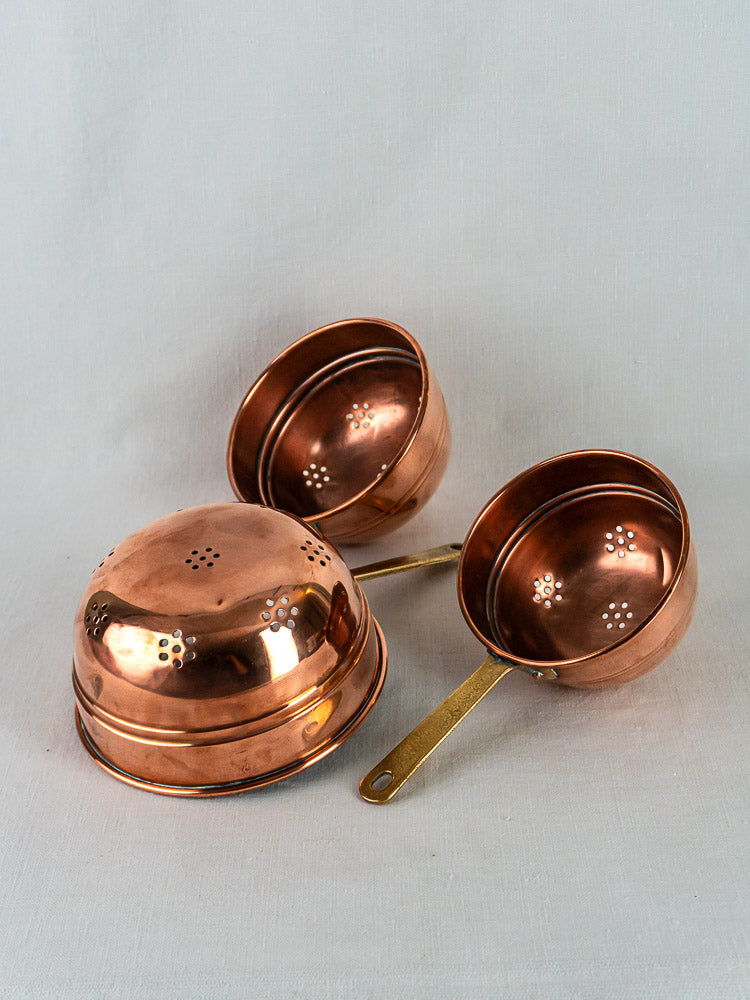 Antique French copper passoire trio with brass handles, flower perforations, on white cloth.