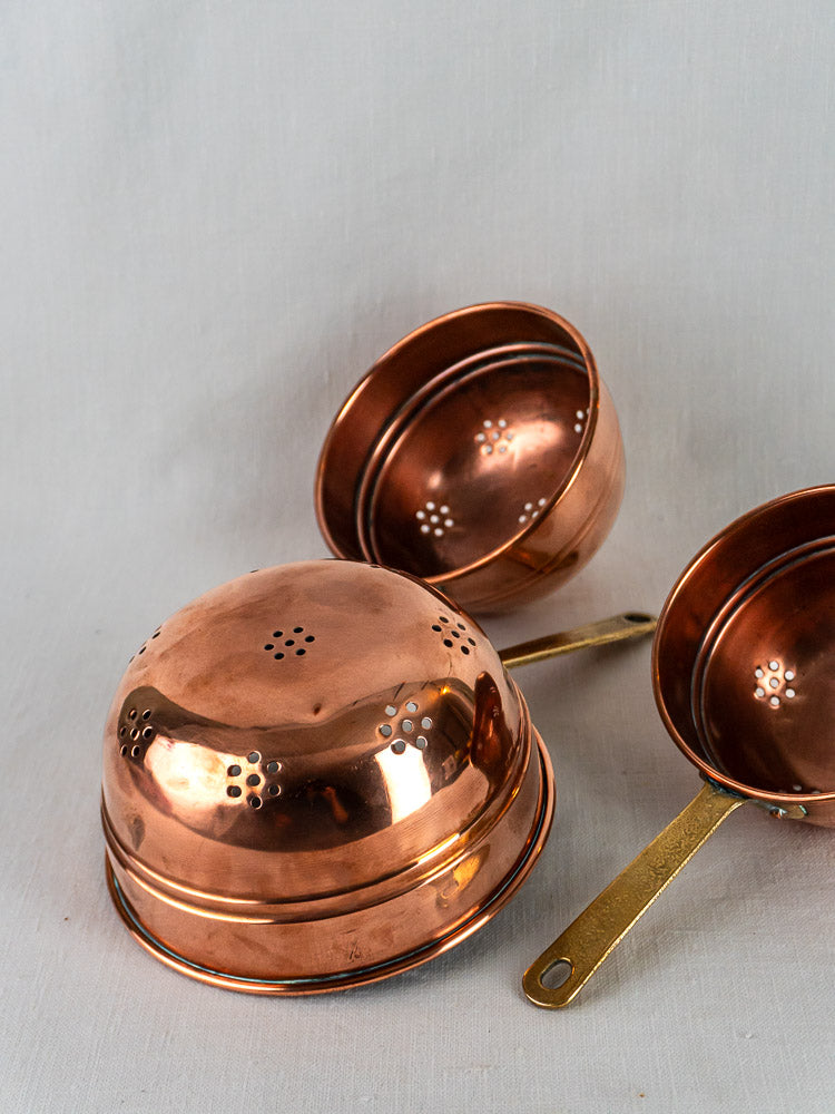 Antique French copper passoires, two upright and one upside down on a light surface.