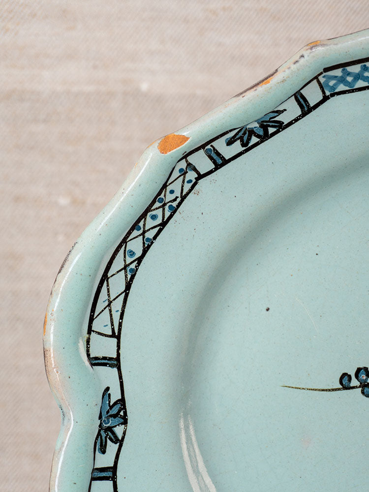 Antique French faience platter, Cul Noir, scalloped edge, black rim, visible chips reveal brown ceramic; on beige textured fabric.