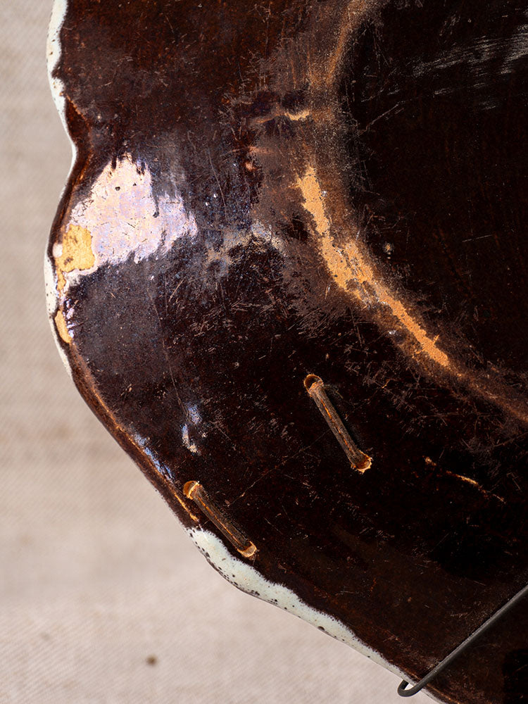 Antique French faience platter with chipped edge, Cul Noir style; partial metal hanger on beige fabric background.