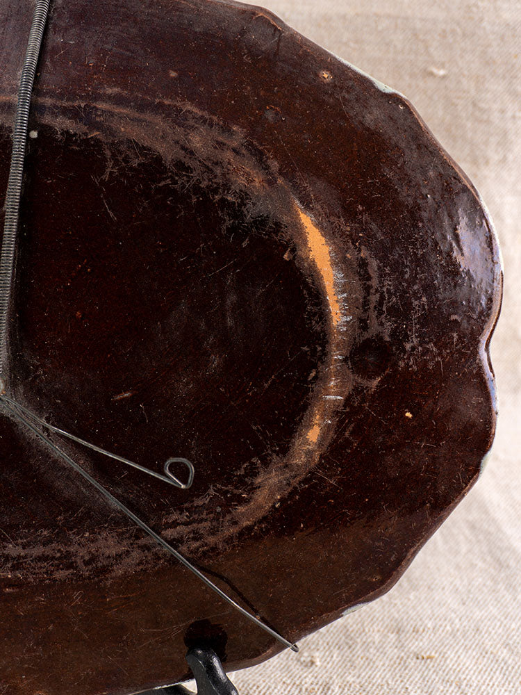 Antique French faience platter underside with metal wire hanger, displaying edge wear and chips on textured beige fabric.