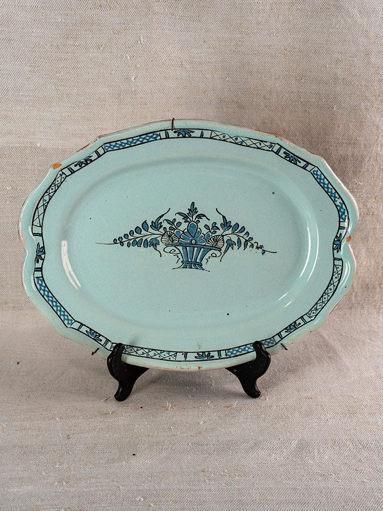 Antique French faience Cul Noir platter with Rouen-style blue floral design, on black stand, neutral fabric backdrop.