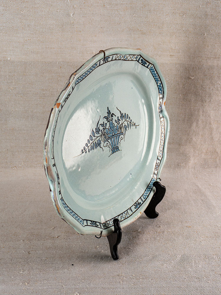 Antique French faience platter with scalloped edge, blue floral vase center, displayed upright on a black stand.