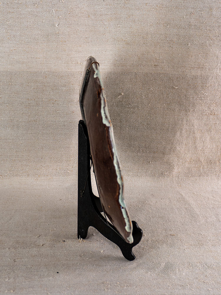 Antique French faience platter, Cul Noir, side view with light rim on black stand, beige fabric backdrop.
