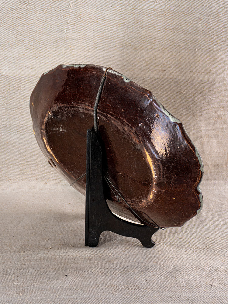 Antique French faience platter with glazed dark brown surface, chipped edges, and metal repair on black stand.