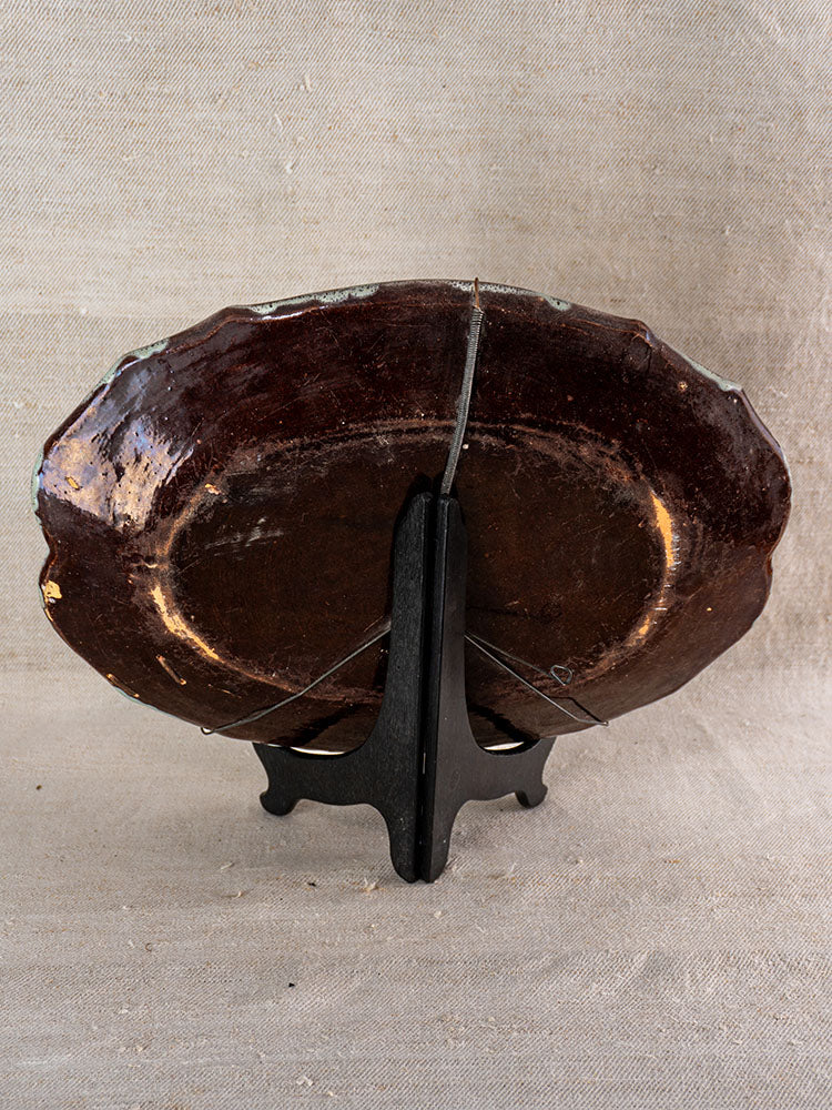 Antique French faience platter with dark brown glaze and wear, upright on black stand, textured beige fabric background.