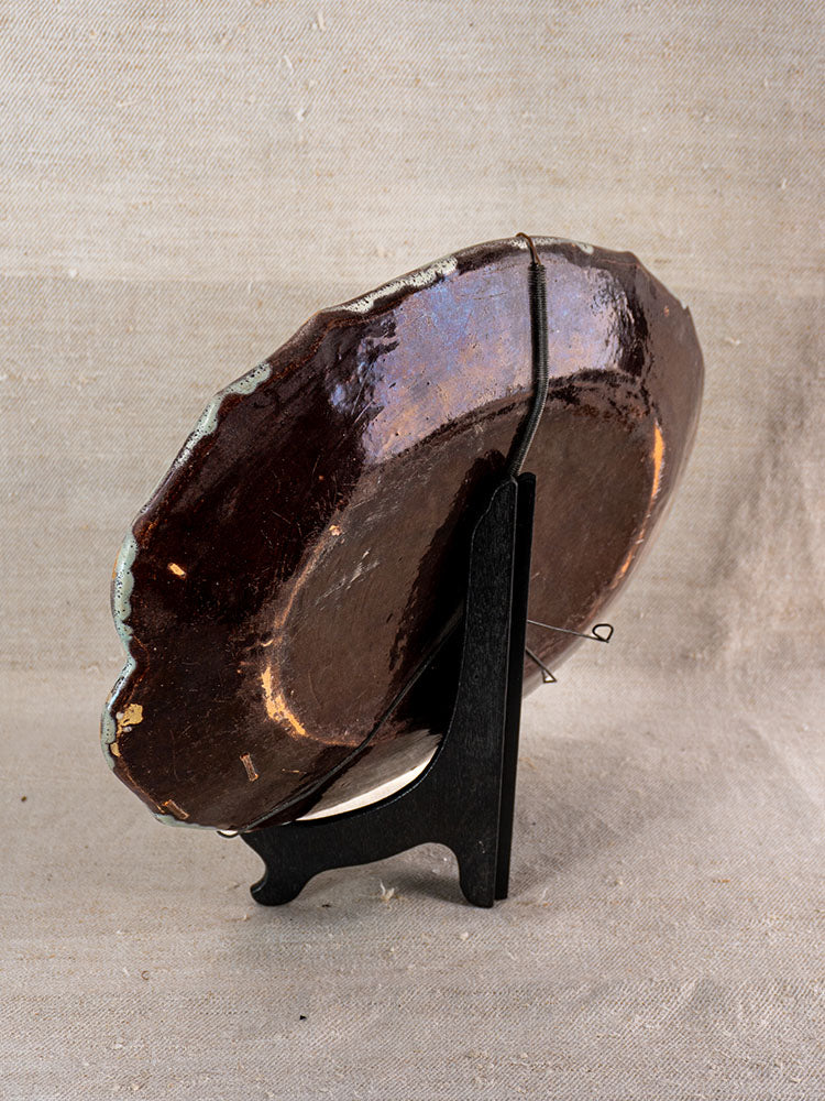 Antique French faience platter Cul Noir, back view on black stand, glossy dark finish, edge wear with chips visible.