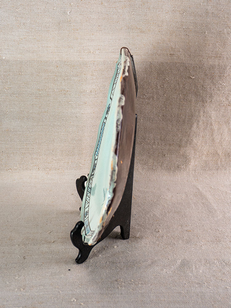 Antique French faience “Cul Noir” platter on black stand, side view, against textured beige fabric backdrop.