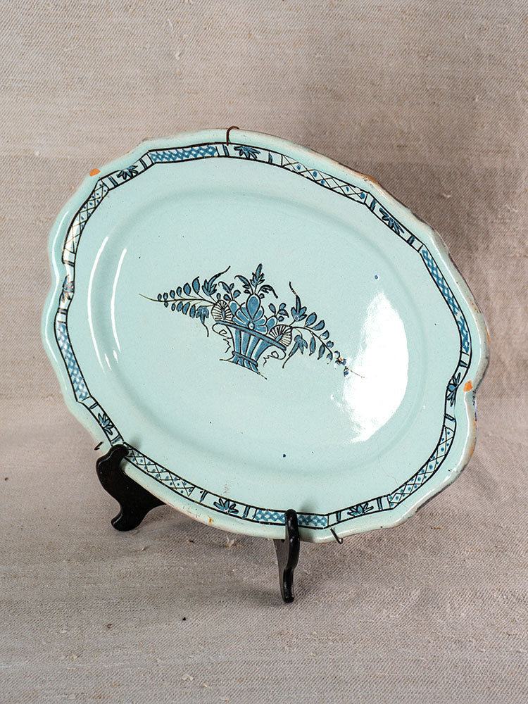Antique French faience platter with pale blue glaze, scalloped edge, floral urn motif, on black stand against beige fabric.