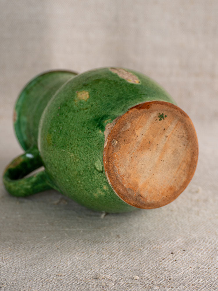 Antique French green glazed earthenware petite cruche with reddish-brown unglazed base on neutral fabric background.