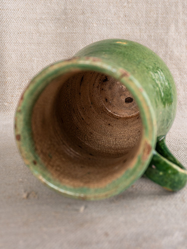 Green glazed French Petite Cruche antique earthenware pitcher with crackled brown interior on beige textured fabric.