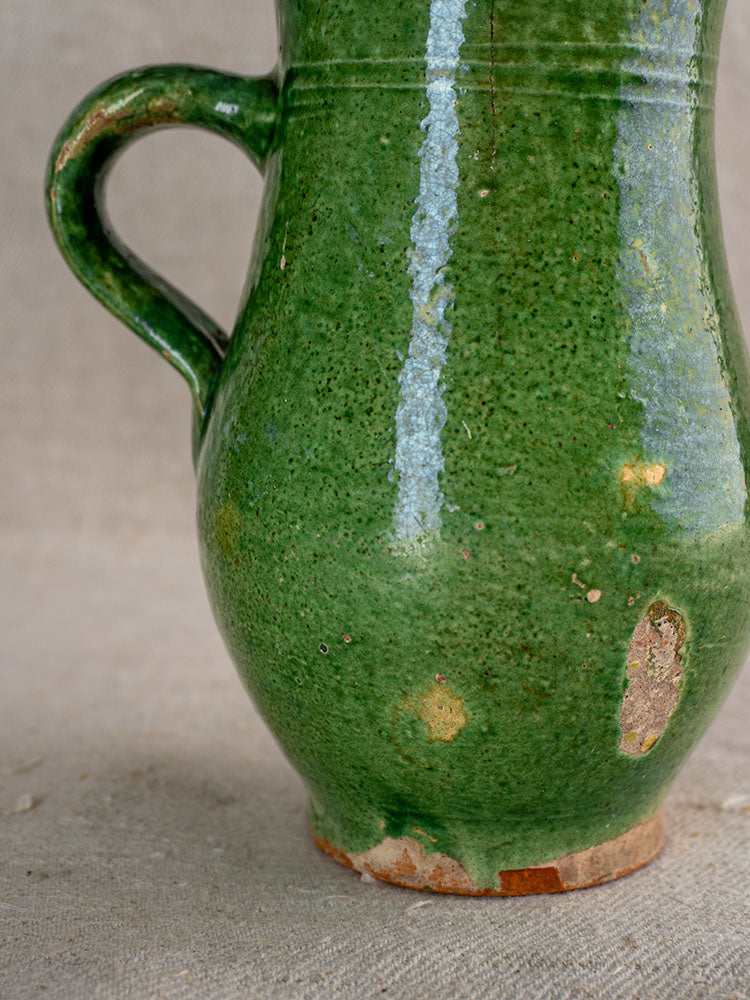 Antique French petite cruche with green glaze, worn and chipped, on a beige textured surface.