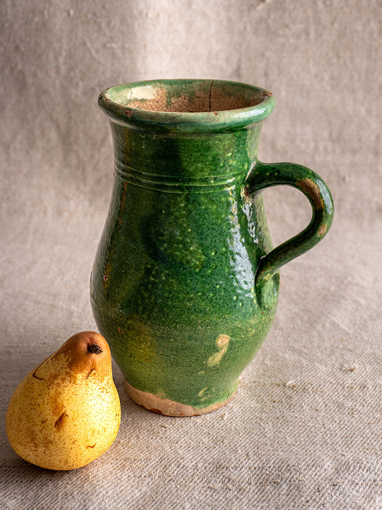 Petite antique French green-glazed earthenware cruche with handle, on beige fabric next to a speckled yellow pear.