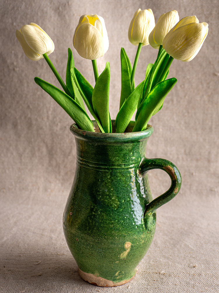 Antique French green-glazed earthenware cruche holding five pale yellow tulips, set against a neutral textured backdrop.
