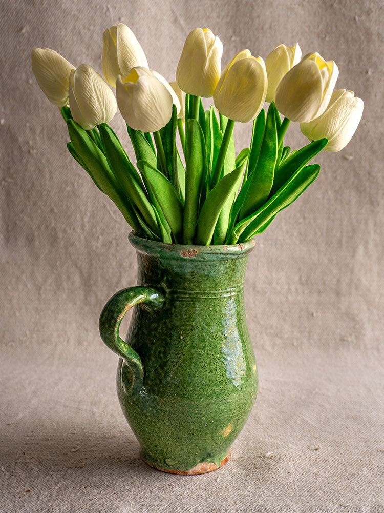 Green glazed French earthenware Petite Cruche pitcher with white tulips, set against a neutral textured background.