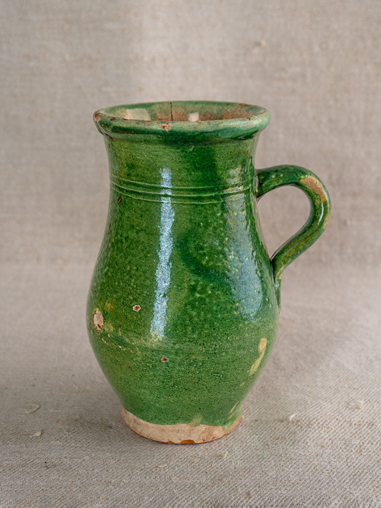 Antique French petite cruche with handle, uneven green glaze, chips, unglazed clay base on beige fabric background.
