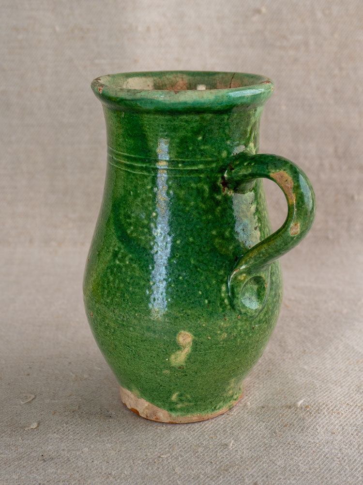 Petite French cruche with green glazed earthenware, terracotta body, curved handle, rustic patina on fabric backdrop.